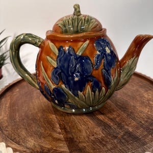 May include: A brown ceramic teapot with a lid decorated with blue and green iris flowers. The teapot has a rounded body and a spout on one side and a handle on the other.