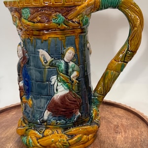 May include: A ceramic pitcher with a detailed design. The pitcher features a dark blue background with figures in relief, a brown and yellow rim, and a twisted handle. The base and handle are adorned with a vine-like pattern.