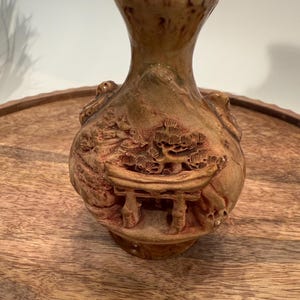 May include: A small, brown ceramic vase with a detailed relief carving of a Japanese torii gate and trees. The vase has a bulbous body and a flared neck. The color is a mix of brown and tan with darker accents.