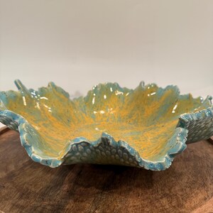 May include: A shallow ceramic bowl with a wavy, leaf-like rim. The interior features a vibrant yellow glaze with speckles, while the exterior and edges are finished in a glossy light blue glaze with a textured, dimpled surface pattern.