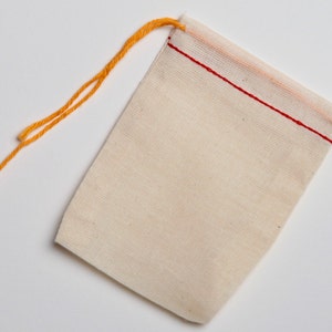 May include: A small, white muslin drawstring bag with a red stitched seam around the top opening. The bag has a yellow drawstring.