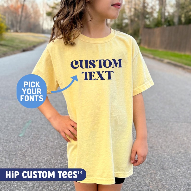 Puede incluir: Una camiseta amarilla con el texto "CUSTOM TEXT" en azul. El texto est&aacute; en un estilo de fuente retro. Un c&iacute;rculo azul con el texto "PICK YOUR FONTS" est&aacute; en la esquina inferior izquierda de la imagen.