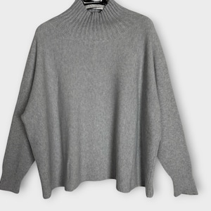 May include: A pale grey jumper with a high, ribbed roll neck and long sleeves. The jumper has a loose fit and ribbed cuffs. The fabric looks soft and comfortable, suitable for cooler weather. The garment is displayed against a plain background.