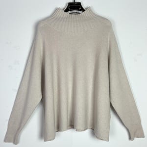 May include: A light beige sweater with a mock neck and long sleeves. The sweater is hanging on a black hanger against a white background. The knitwear has a ribbed collar and cuffs.
