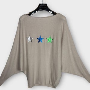 Op de afbeelding: Een beige trui met een boothals en lange mouwen, hangend aan een zwarte hanger. De trui heeft drie stervormige versieringen in zilver, blauw en groen over de borst.