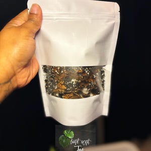 May include: A white, resealable pouch with a clear window, held by a hand. The window reveals a dark, textured substance. The pouch has the text "planted with Jae" and a graphic of green leaves.