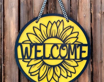 May include: A circular wooden door sign featuring a bright yellow sunflower design with a black border. The word WELCOME is cut out in black lettering across the center of the sunflower. A black and white gingham ribbon is attached for hanging.