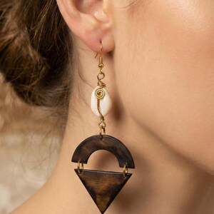 May include: A pair of dangle earrings featuring a white seashell, gold-colored wire, and dark brown wooden geometric shapes. The earrings have a hook closure and are designed for pierced ears. The earrings are a unique accessory.