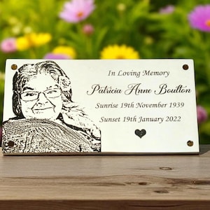 May include: A rectangular wooden memorial plaque with a black and white portrait of a woman wearing glasses. The plaque includes the text "In Loving Memory" and the name "Patricia Anne Boulton" along with birth and death dates.