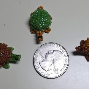 May include: Three beaded turtle figurines in shades of brown, green, and tan are displayed next to a U.S. quarter. The turtles are crafted with small beads, showcasing detailed shell and body designs. The quarter provides a size reference.