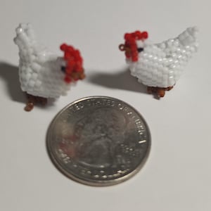 May include: Two handmade beaded chicken figurines. The chickens are white with red combs and brown feet. A U.S. quarter is next to the chickens for size comparison. The chickens are likely earrings or small decorative items.