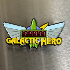 May include: A colorful Galactic Hero pin with a digital display showing "999999". The pin features a green, blue, and yellow design with wing accents. The text "GALACTIC HERO" is in yellow with a black outline.