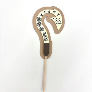 May include: A brown cardboard cutout of a hook with a white design, including waves, spirals, and arrows. The hook is attached to a wooden stick.