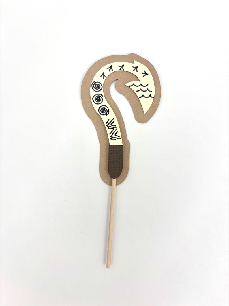 May include: A brown cardboard cutout of a hook with a white design, including waves, spirals, and arrows. The hook is attached to a wooden stick.