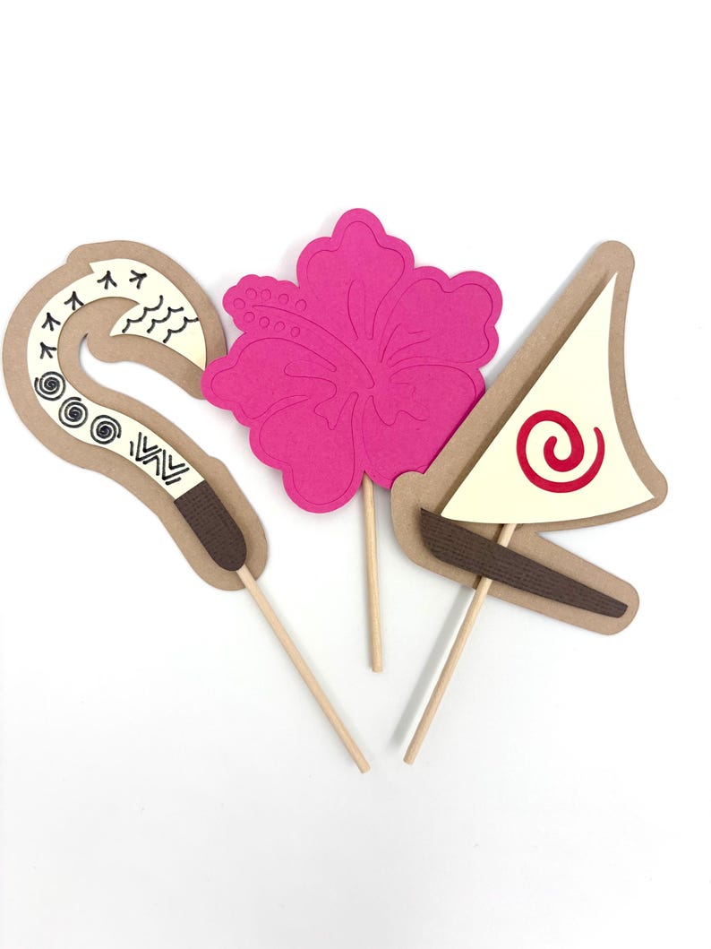 May include: Three paper cupcake toppers featuring a pink hibiscus flower, a brown and white hook, and a white sailboat with a red spiral design.