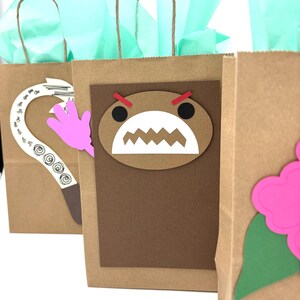 May include: Three brown paper gift bags with decorative cutouts. One bag features a Domo-kun face, another a pink hand, and the third a pink flower. Each bag has a twisted paper handle and is filled with teal tissue paper.