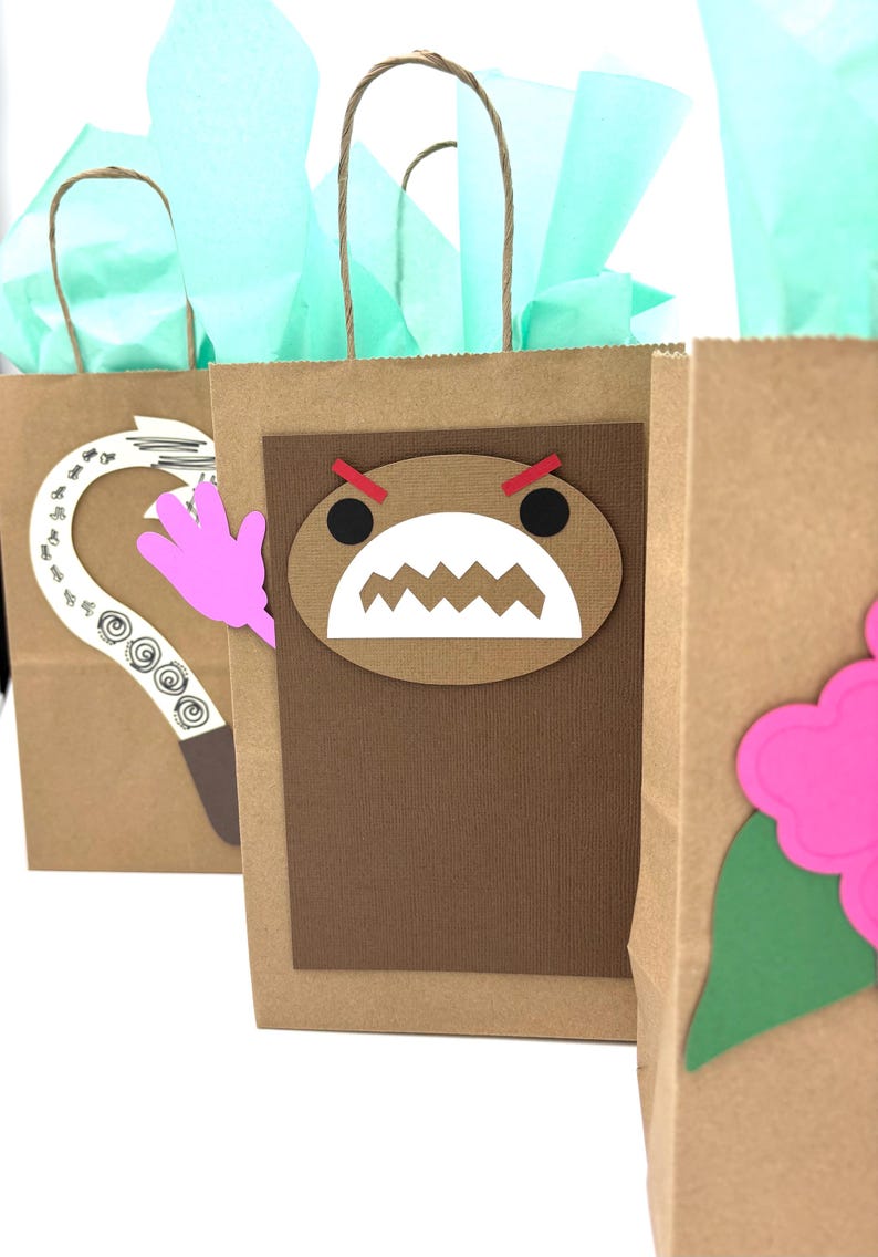 May include: Three brown paper gift bags with decorative cutouts. One bag features a Domo-kun face, another a pink hand, and the third a pink flower. Each bag has a twisted paper handle and is filled with teal tissue paper.