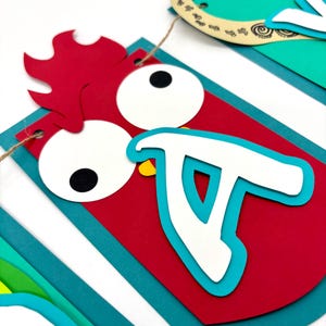 May include: A colourful paper banner with the word "Aloha" spelled out in white letters. Each letter is decorated with a different animal or design. The "A" is a red rooster with a yellow beak and large eyes. The "L" is a green palm tree. The "O" is a blue fish. The "H" is a brown horseshoe. The "A" is a green pineapple.