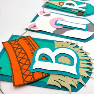 May include: A colourful paper banner with the word "Birthday" spelled out in white letters. Each letter is layered with different coloured paper and has a farm animal theme. The banner is strung together with twine.
