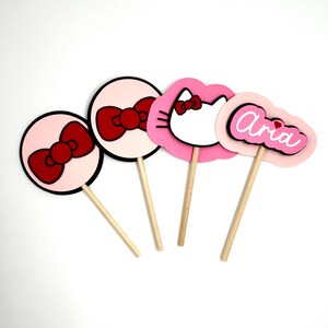 Może przedstawiać: Cztery różowe i czerwone ozdoby do babeczek z motywem Hello Kitty. Ozdoby przedstawiają czerwoną kokardkę, twarz Hello Kitty i imię "Aria" w różowych literach.