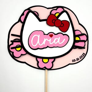 Może przedstawiać: Różowy i biały topper na tort w stylu Hello Kitty z imieniem "Aria" i datą "03-31-2025" napisanymi na nim.