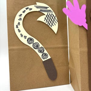 May include: Brown paper gift bag with a decorative cream-colored hook and a pink hand cutout. The hook features black line art and spiral designs. The bag contains teal tissue paper.