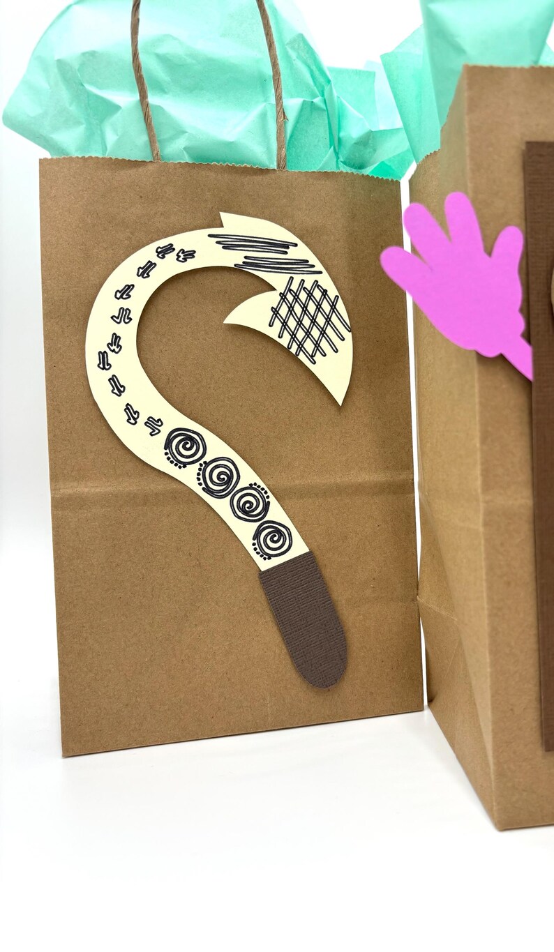 May include: Brown paper gift bag with a decorative cream-colored hook and a pink hand cutout. The hook features black line art and spiral designs. The bag contains teal tissue paper.