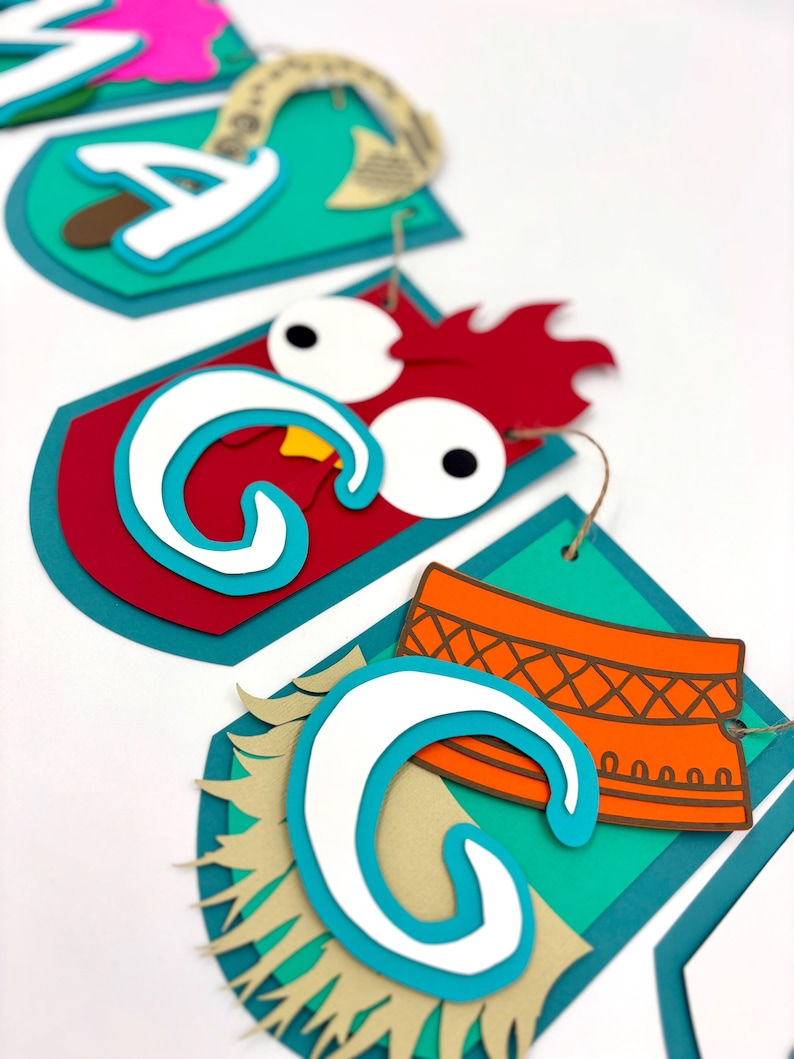 May include: A colourful paper banner with the word "Moana" spelled out in large, white letters. Each letter is decorated with a different design, including a rooster, a flower, and a coconut.
