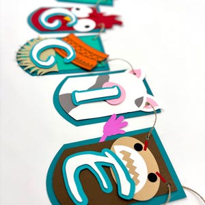May include: A colourful paper banner with animal characters and the letters "G I Z E L L E" in white letters on a turquoise background. The characters include a rooster, a cow, a pig, and a monster.