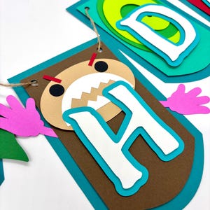 May include: A colourful paper banner with the letters "Happy" in a monster theme. The letter "H" is brown with a white letter and a monster face with red eyes and teeth.