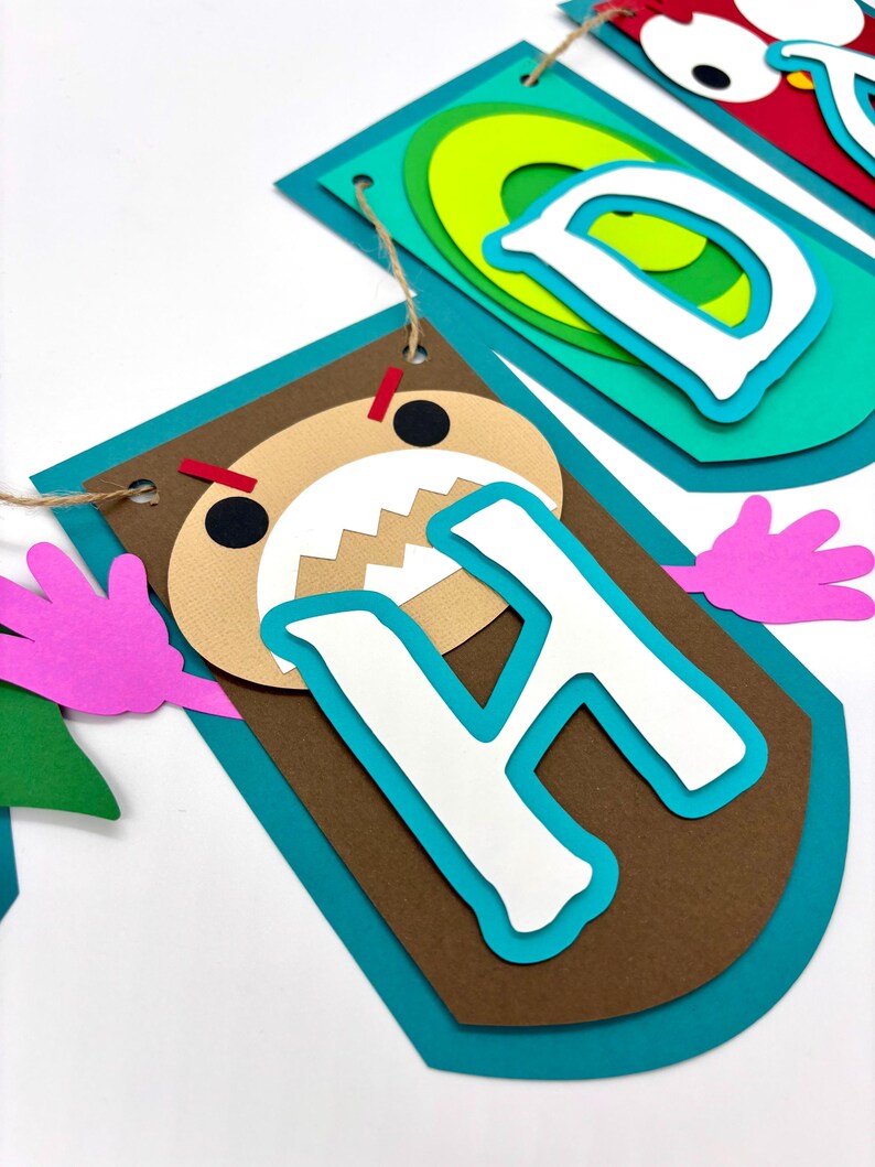 May include: A colourful paper banner with the letters "Happy" in a monster theme. The letter "H" is brown with a white letter and a monster face with red eyes and teeth.