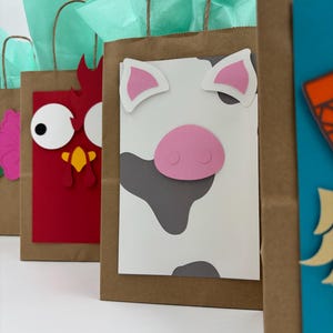 May include: Brown paper gift bags decorated with farm animal faces, including a cow, chicken, and pig. Each bag has a different animal face made of colorful paper cutouts. The bags are filled with teal tissue paper and have brown paper handles.