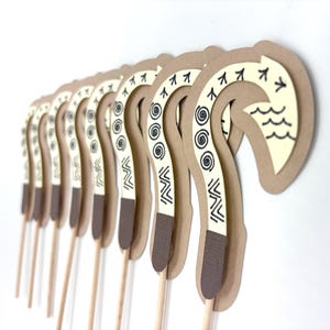 May include: A set of 10 brown and white paper cupcake toppers shaped like a traditional Hawaiian hook, or "hei", with black and white geometric patterns. The toppers are attached to wooden skewers.