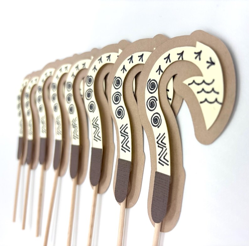 May include: A set of 10 brown and white paper cupcake toppers shaped like a traditional Hawaiian hook, or "hei", with black and white geometric patterns. The toppers are attached to wooden skewers.