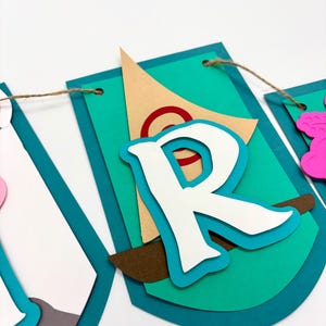 May include: A teal blue banner with a white letter "R" on it. The letter is layered on top of a brown sailboat with a red circle on the sail. The banner is strung with twine.
