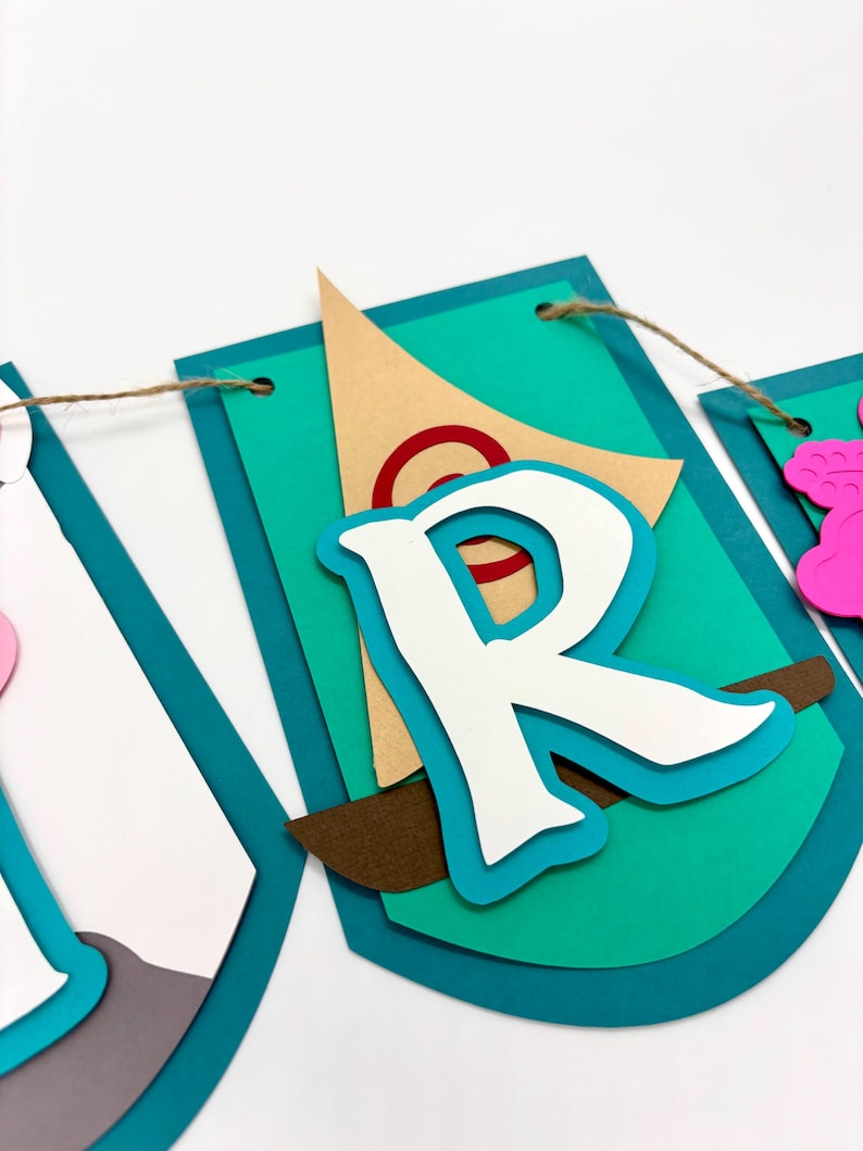 May include: A teal blue banner with a white letter "R" on it. The letter is layered on top of a brown sailboat with a red circle on the sail. The banner is strung with twine.