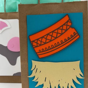 May include: Brown paper gift bags with teal tissue paper and brown twine handles. One bag has a blue rectangle with an orange design and a tan cutout. Another bag has a cow face design.