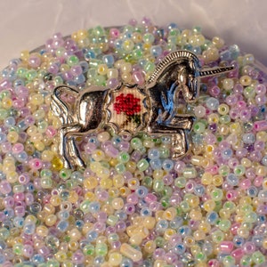 Könnte beinhalten: Eine silberne Einhorn-Figur mit einer roten, gestickten Blume auf dem Rücken, umgeben von schillernden pastellfarbenen Perlen.