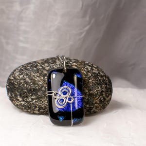 May include: A rectangular pendant with a black and blue design, accented with silver wire detailing. The pendant hangs from a silver chain. The blue colour is concentrated in the centre of the pendant. The pendant is resting on a dark grey rock.