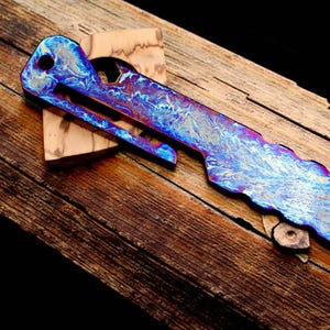 May include: A multi-tool knife with a vibrant blue and purple marbled pattern. The knife is set on a wooden surface, showcasing its unique design and craftsmanship. The blade has a distinctive shape, and the handle features a pocket clip.