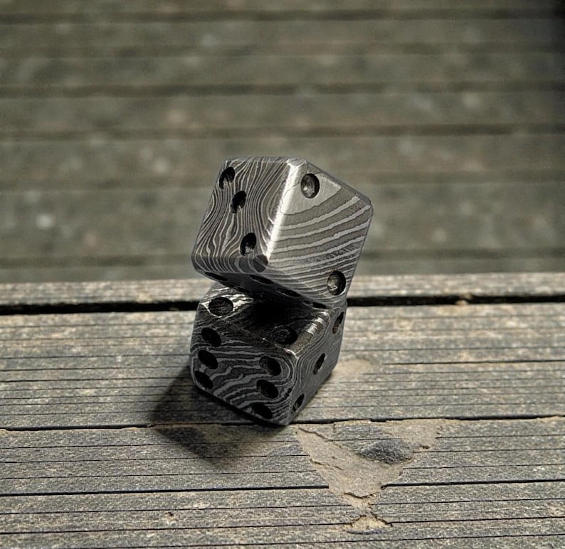 Peut inclure: Deux d&eacute;s en m&eacute;tal avec un motif unique, l'un reposant sur l'autre. Les d&eacute;s sont gris fonc&eacute; avec une surface textur&eacute;e et des points noirs repr&eacute;sentant les chiffres. Ils sont pos&eacute;s sur une surface en bois.