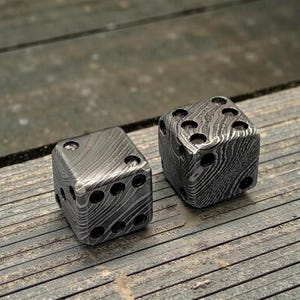Peut inclure: Deux d&eacute;s en m&eacute;tal avec un motif unique. Chaque d&eacute; comporte des points noirs repr&eacute;sentant les chiffres. Les d&eacute;s sont en m&eacute;tal fonc&eacute; et textur&eacute;, pr&eacute;sent&eacute;s sur une surface en bois.