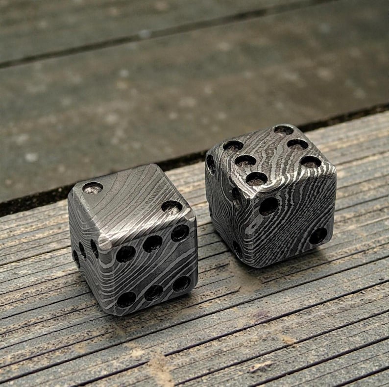 Peut inclure: Deux d&eacute;s en m&eacute;tal avec un motif unique. Chaque d&eacute; comporte des points noirs repr&eacute;sentant les chiffres. Les d&eacute;s sont en m&eacute;tal fonc&eacute; et textur&eacute;, pr&eacute;sent&eacute;s sur une surface en bois.