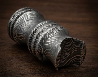 Peut inclure: Un objet cylindrique en métal présentant un motif en couches ondulées distinctes dans des tons de gris, noir et argent. La surface affiche une texture complexe et fluide avec une série de crêtes horizontales et une extrémité concave.