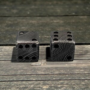 Peut inclure: Deux d&eacute;s en m&eacute;tal avec un motif unique. Les d&eacute;s sont gris fonc&eacute; avec une texture tourbillonnante semblable &agrave; du grain de bois. Chaque d&eacute; comporte le nombre standard d'indentations circulaires, repr&eacute;sentant les chiffres un &agrave; six.
