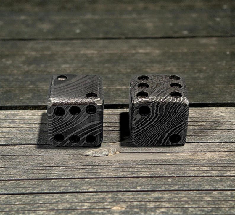 Peut inclure: Deux d&eacute;s en m&eacute;tal avec un motif unique. Les d&eacute;s sont gris fonc&eacute; avec une texture tourbillonnante semblable &agrave; du grain de bois. Chaque d&eacute; comporte le nombre standard d'indentations circulaires, repr&eacute;sentant les chiffres un &agrave; six.