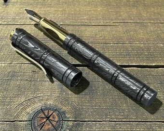Peut inclure: Un stylo plume de luxe avec un motif de grain de bois noir et gris. Le stylo a une plume et des accents dorés, et le capuchon est détaché. Le stylo est posé sur une surface en bois. Le design du stylo rappelle un baril.