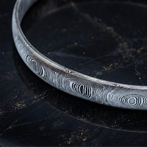 May include: A circular bangle bracelet featuring a distinct Damascus steel pattern with alternating black and silver-white wavy, swirling lines that wrap continuously around the entire surface of the metal band, creating a high-contrast visual texture.
