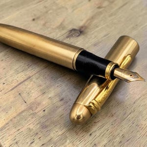Op de afbeelding: Een goudkleurige vulpen met een geborstelde metalen afwerking op de houder en een gepolijste dop. De pen heeft een zwart gripgedeelte en een metalen penpunt. De dop is voorzien van een gouden clip en ligt naast de houder van de pen.
