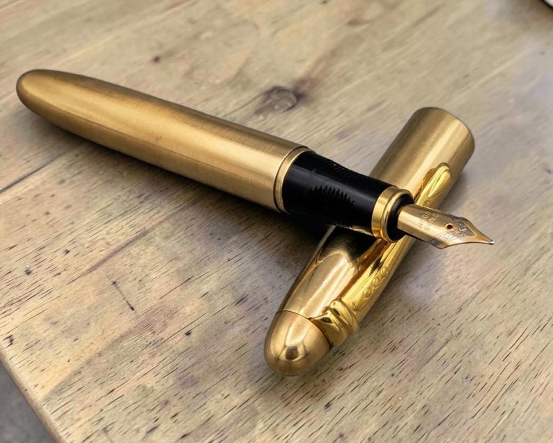 Op de afbeelding: Een goudkleurige vulpen met een geborstelde metalen afwerking op de houder en een gepolijste dop. De pen heeft een zwart gripgedeelte en een metalen penpunt. De dop is voorzien van een gouden clip en ligt naast de houder van de pen.